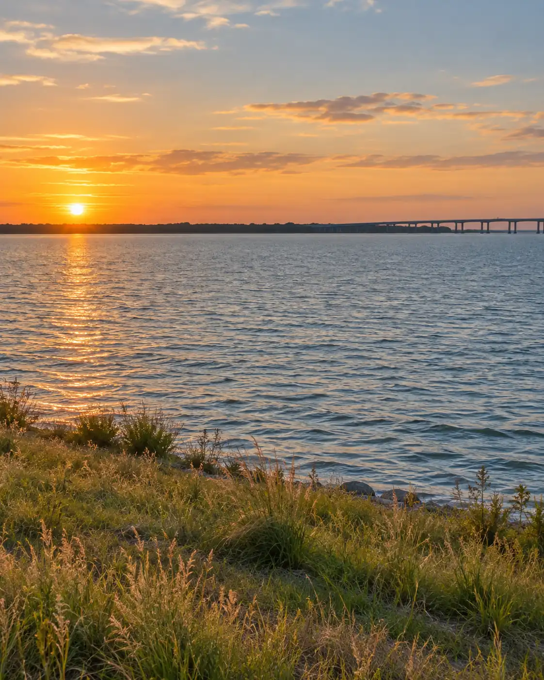 Illustrative reservoir shoreline with calm water and a distant bridge at sunset